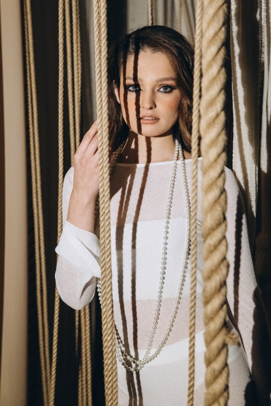 Портрет женщины в белой одежде с длинным ожерельем среди декоративных верёвок с драматичным освещением
