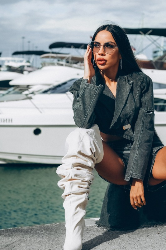 Девушка в белых сапогах и джинсовой куртке позирует у причала с яхтами, разговаривая по телефону в солнечных очках.