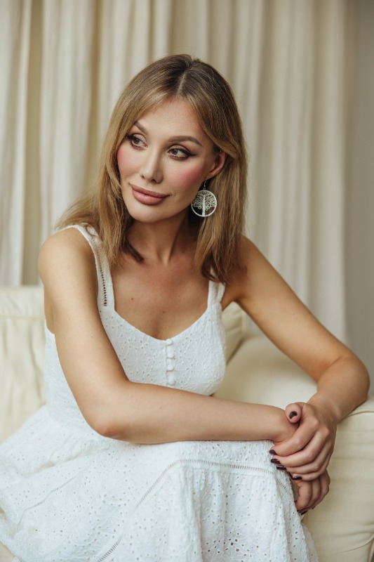 Женщина в белом платье с серьгами сидит на светлом диване на фоне бежевых штор