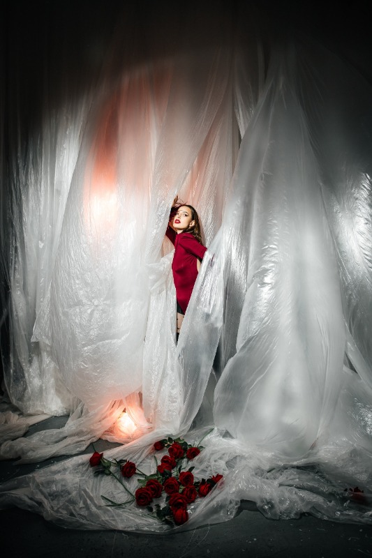 Женщина в красном платье стоит среди полупрозрачных белых занавесей, у её ног лежат красные розы и горит свеча.
