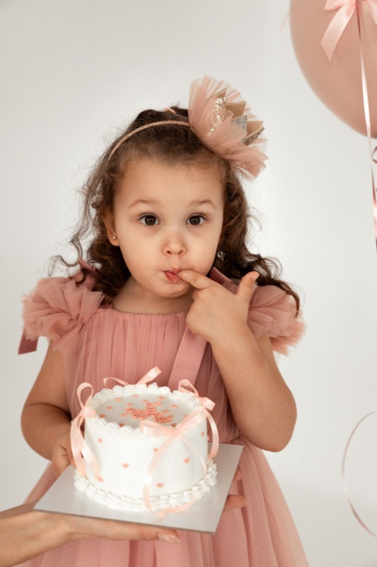 Маленькая девочка в розовом платье с бантом держит праздничный торт и облизывает палец с кремом