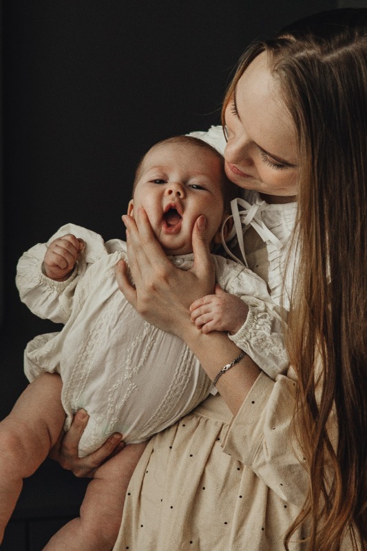 Мама в светлом платье держит младенца в белой одежде на тёмном фоне, малыш зевает