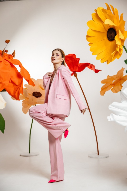 Женщина в розовом костюме позирует среди больших бумажных цветов на белом фоне в студии
