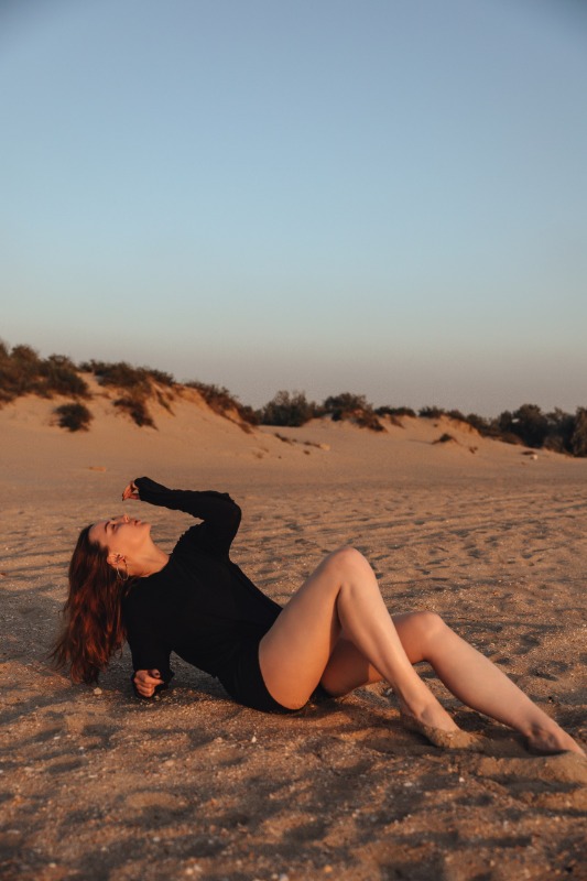 Девушка в черном боди лежит на песке в лучах заката с дюнами и кустарником на фоне