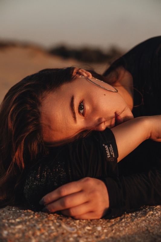 Девушка лежит на песке в лучах заходящего солнца, смотрит в камеру, одета в чёрную футболку