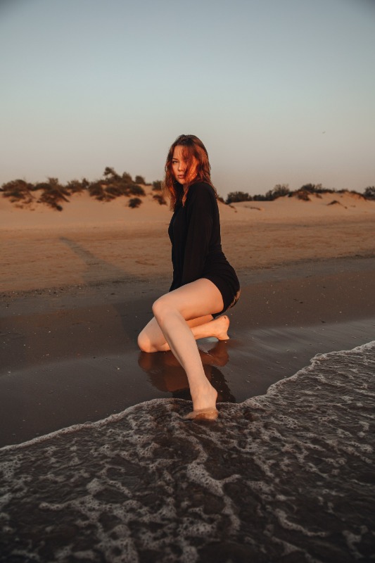 Девушка в черной одежде сидит на камне у кромки воды на пляже во время заката