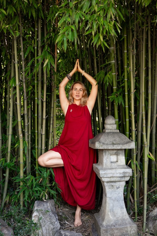Женщина в красном платье выполняет позу йоги на одной ноге на фоне бамбуковой рощи и каменного фонаря