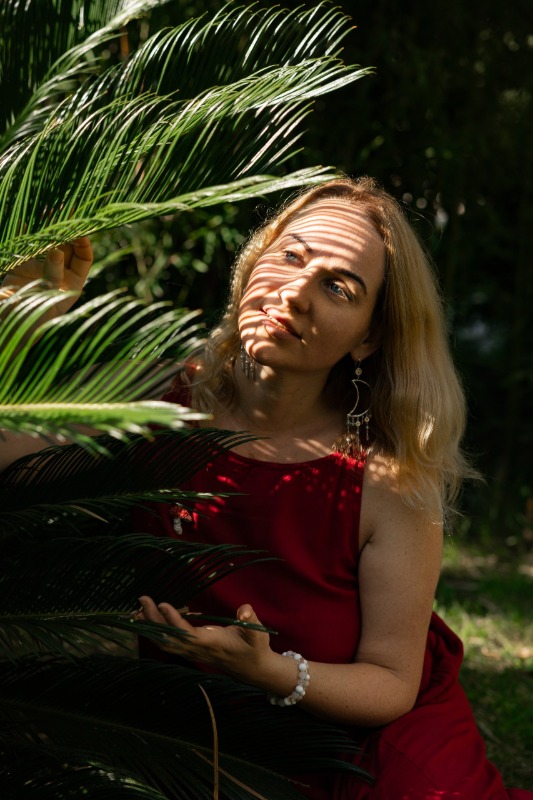Женщина в красном платье среди пальмовых листьев, солнечный свет создает тени на лице