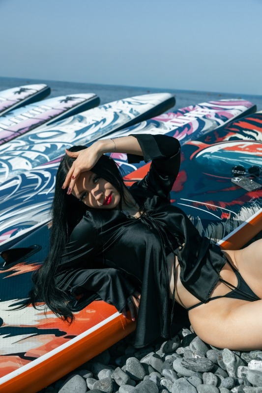 Девушка в черном купальнике лежит на цветных досках для серфинга на галечном пляже у моря