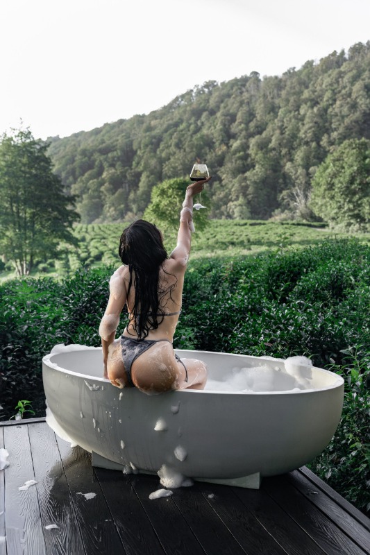Женщина в ванне на открытом воздухе с бокалом вина на фоне виноградников и холмов